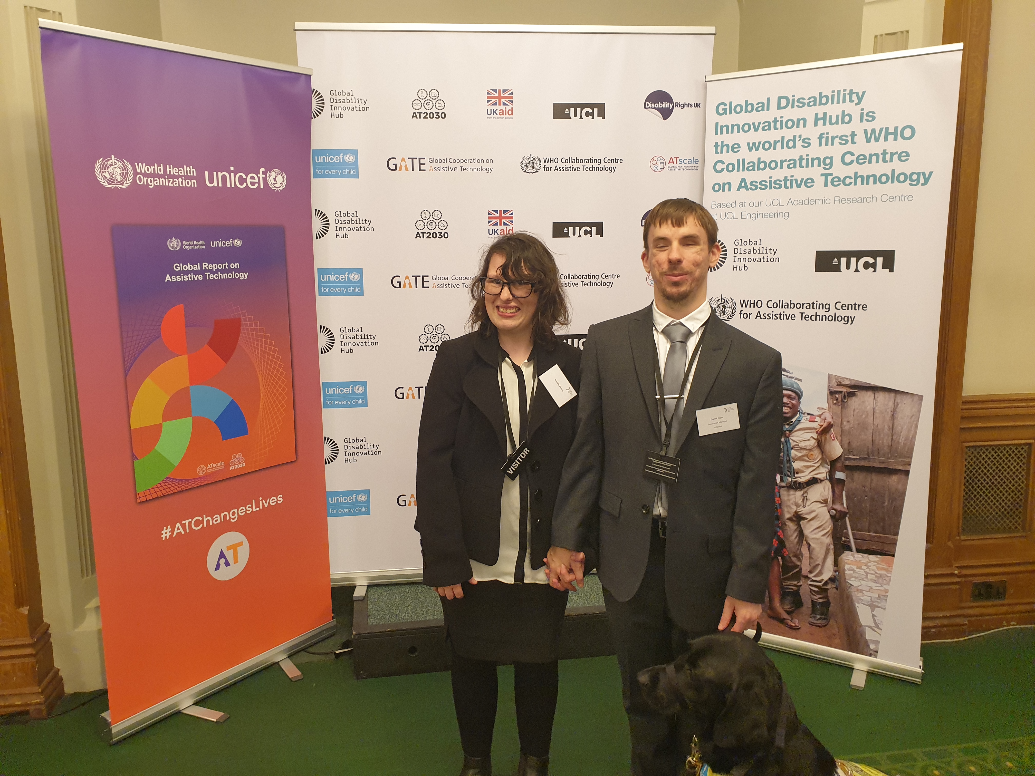 Daniel and Danielle are photographed in the UK Parliament. They are wearing smart clothing. A guide dog is sitting in front of them. In the background multiple signs are visible, such as logos of University College London, World Health Organisation, and GDI Hub. Other signs read 'AT changes lives' and 'Launching the Global Report on Assistive Technology'.