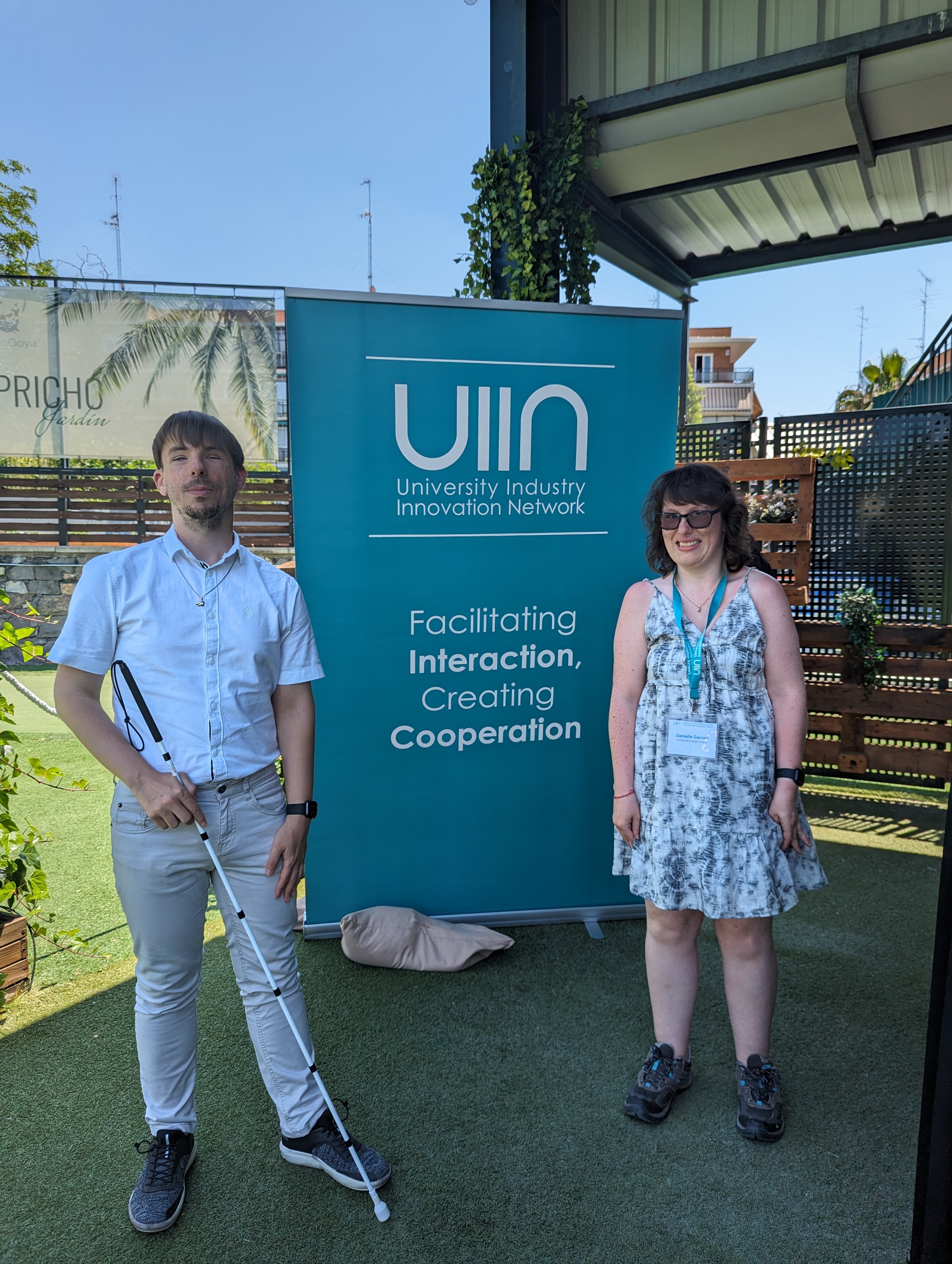 Daniel and Danielle are dressed in smart clothes in an outdoor setting, in front of a board that says 'University Industry Innovation network, Facilitating interaction and creating cooperation'.