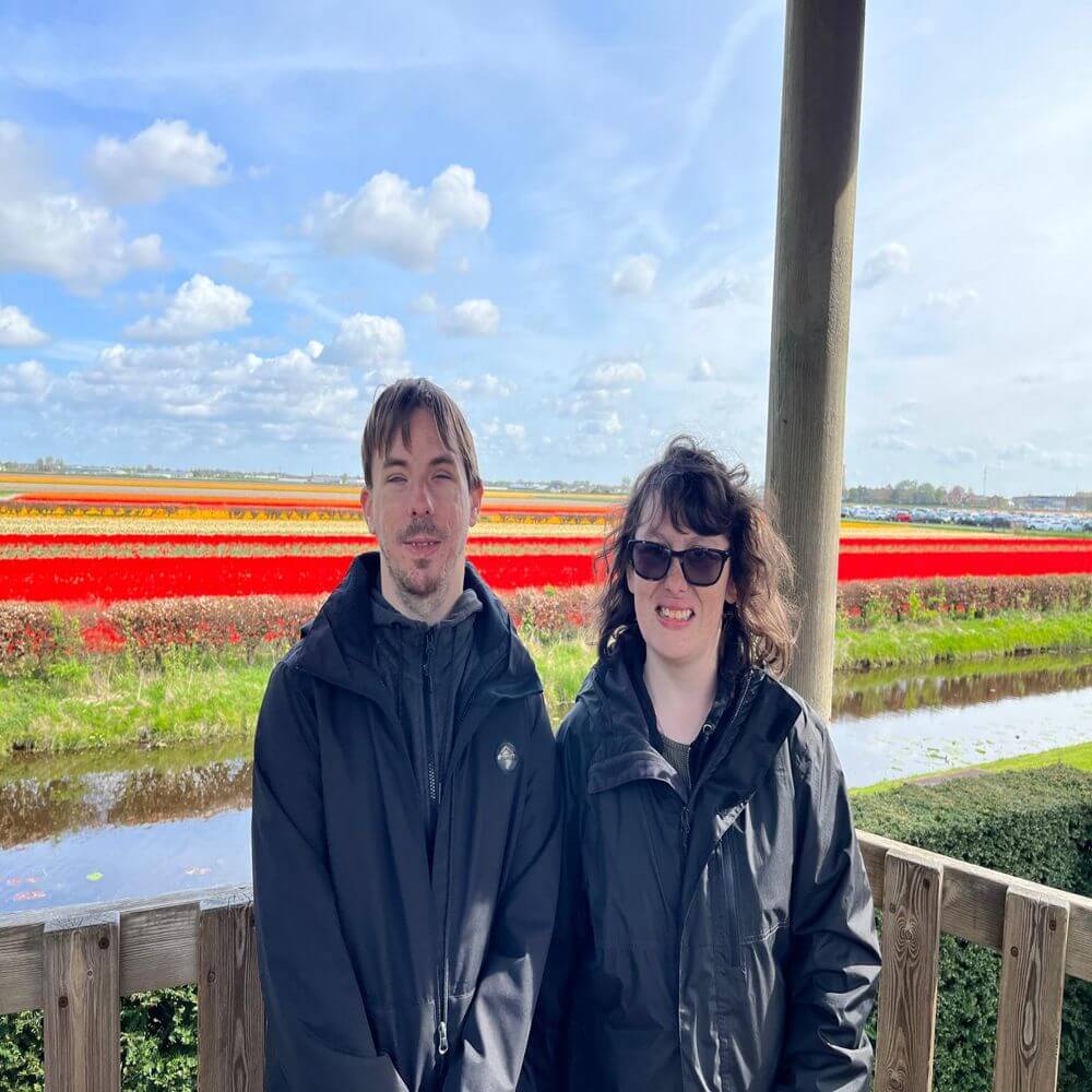 Daniel and Danielle are standing on a platform, with vast fields of colourful tulips and a canal in the background. Both of them smile at the camera.