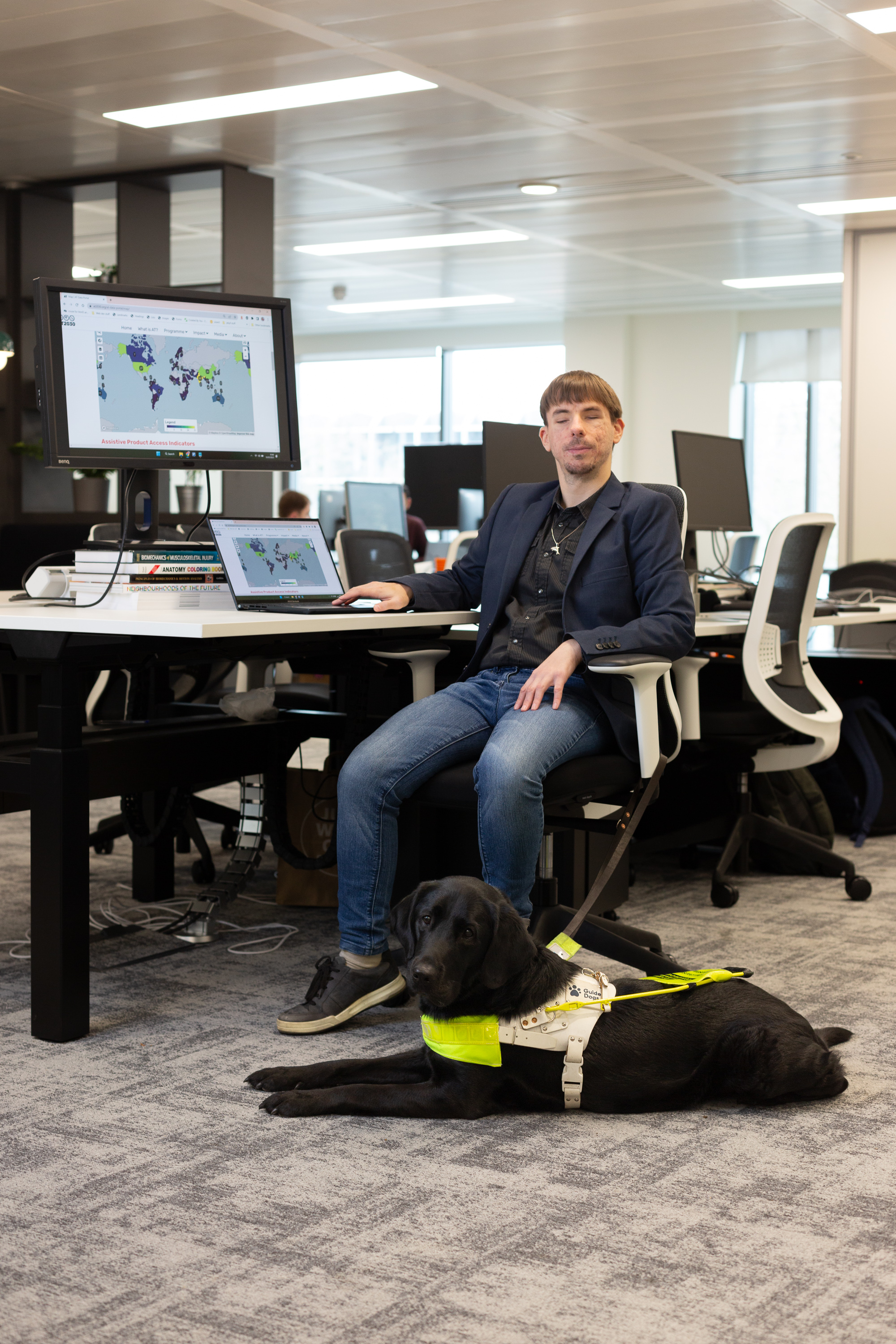 Daniel is wearing a dark blue suit jacket and blue jeans he is sitting at a table with a computer next to him and his black guide dog Anna laying beside him they are both facing the camera.